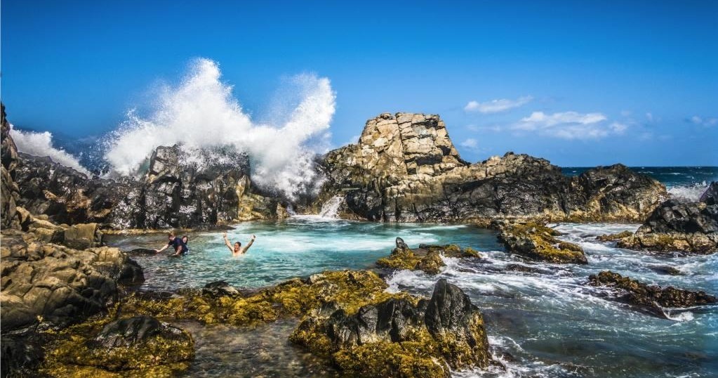 Waves crashing on rocky shore with people swimming in a natural pool.