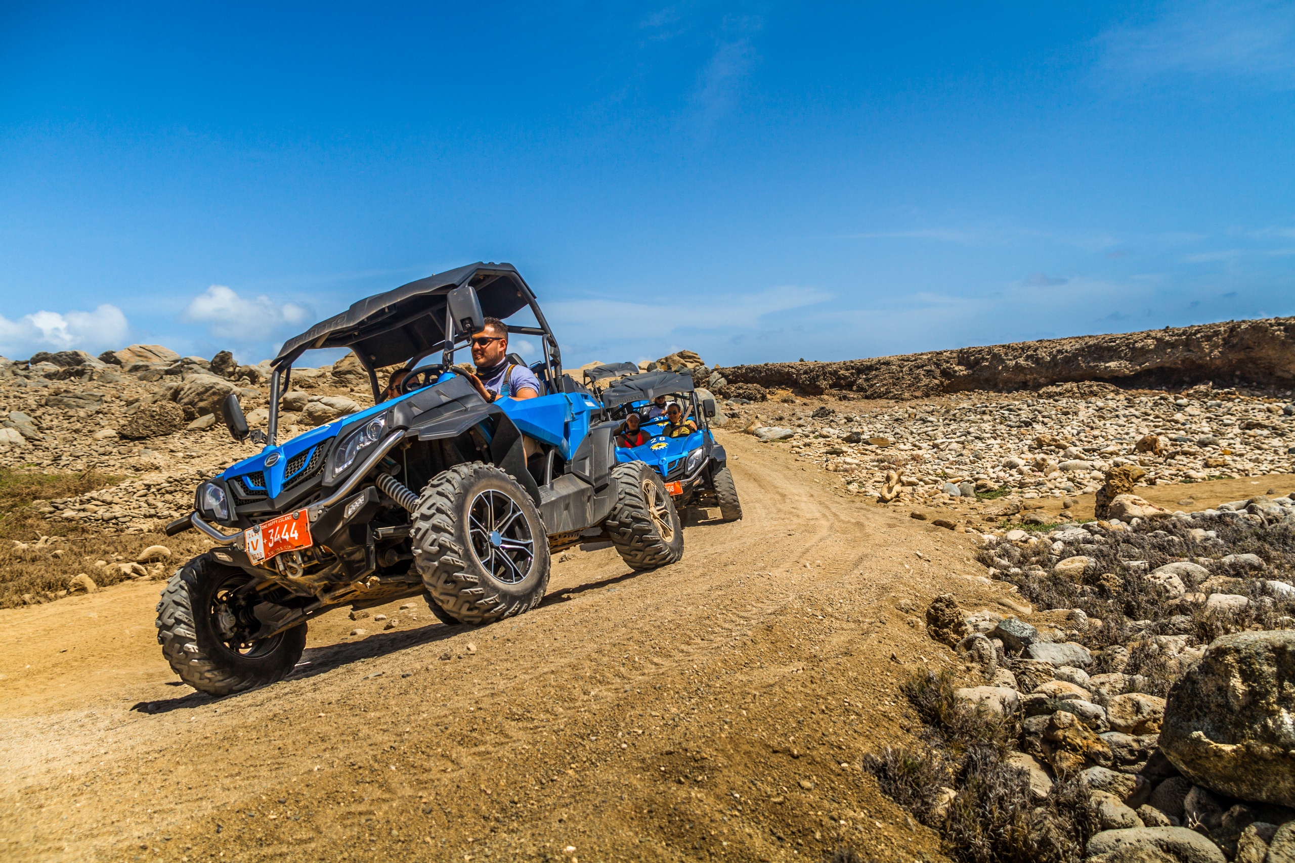 Two off-road vehicles driving on a rocky desert trail under a blue sky.