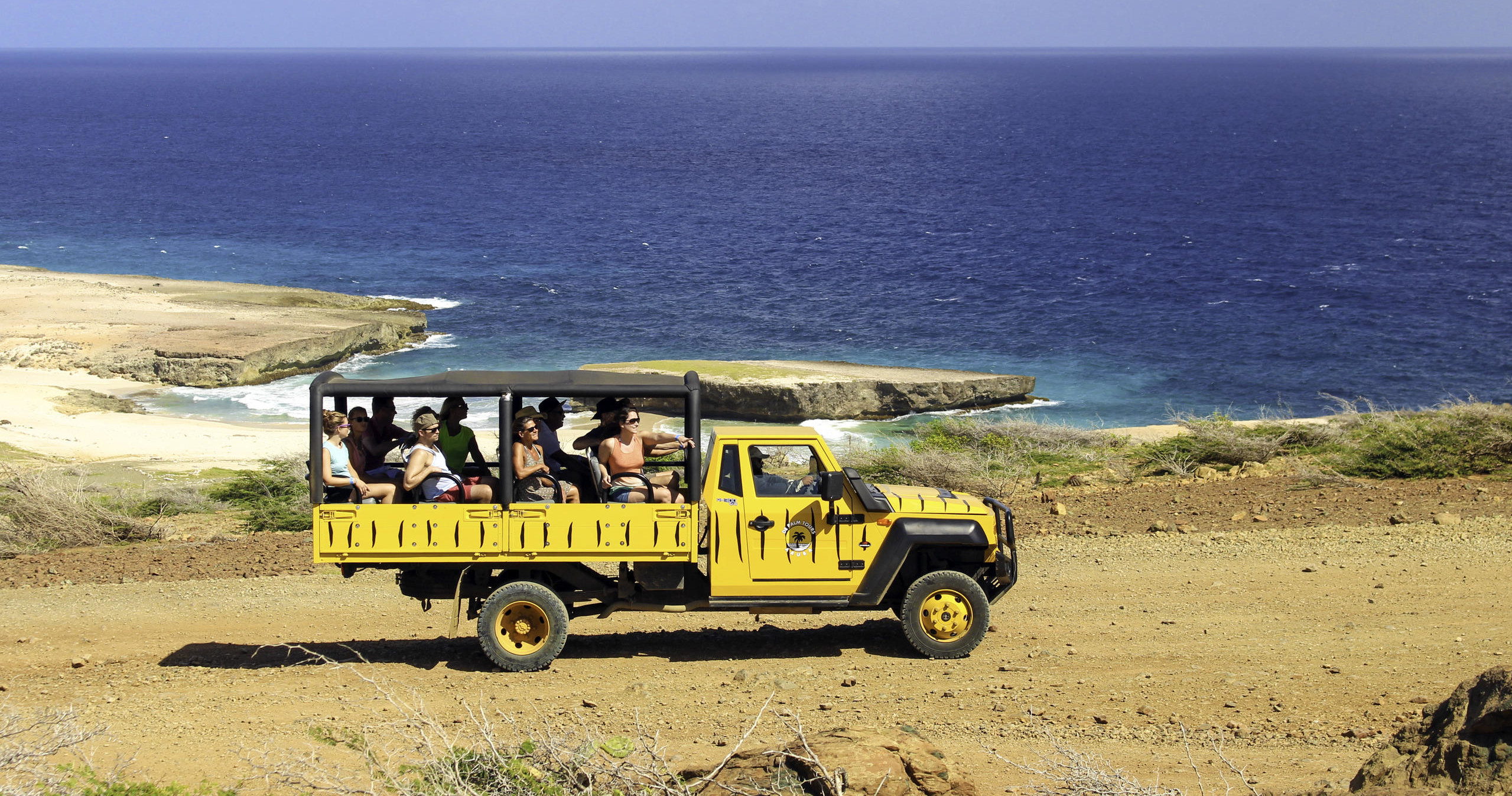 Tourists in yellow safari jeep driving on dirt road near ocean.