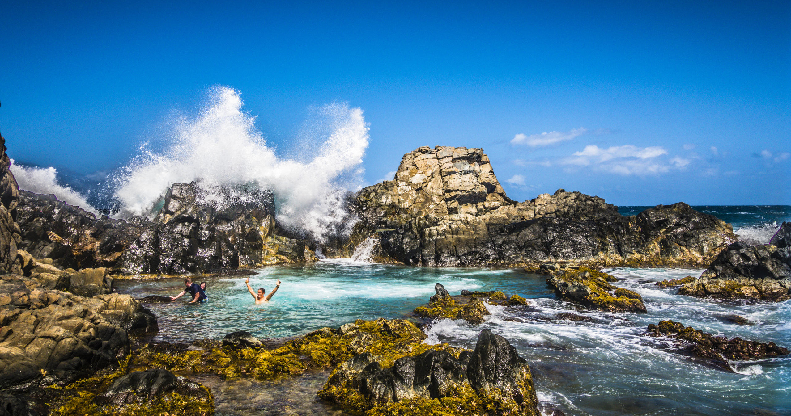 Waves crash on rocks near a natural pool with people swimming.