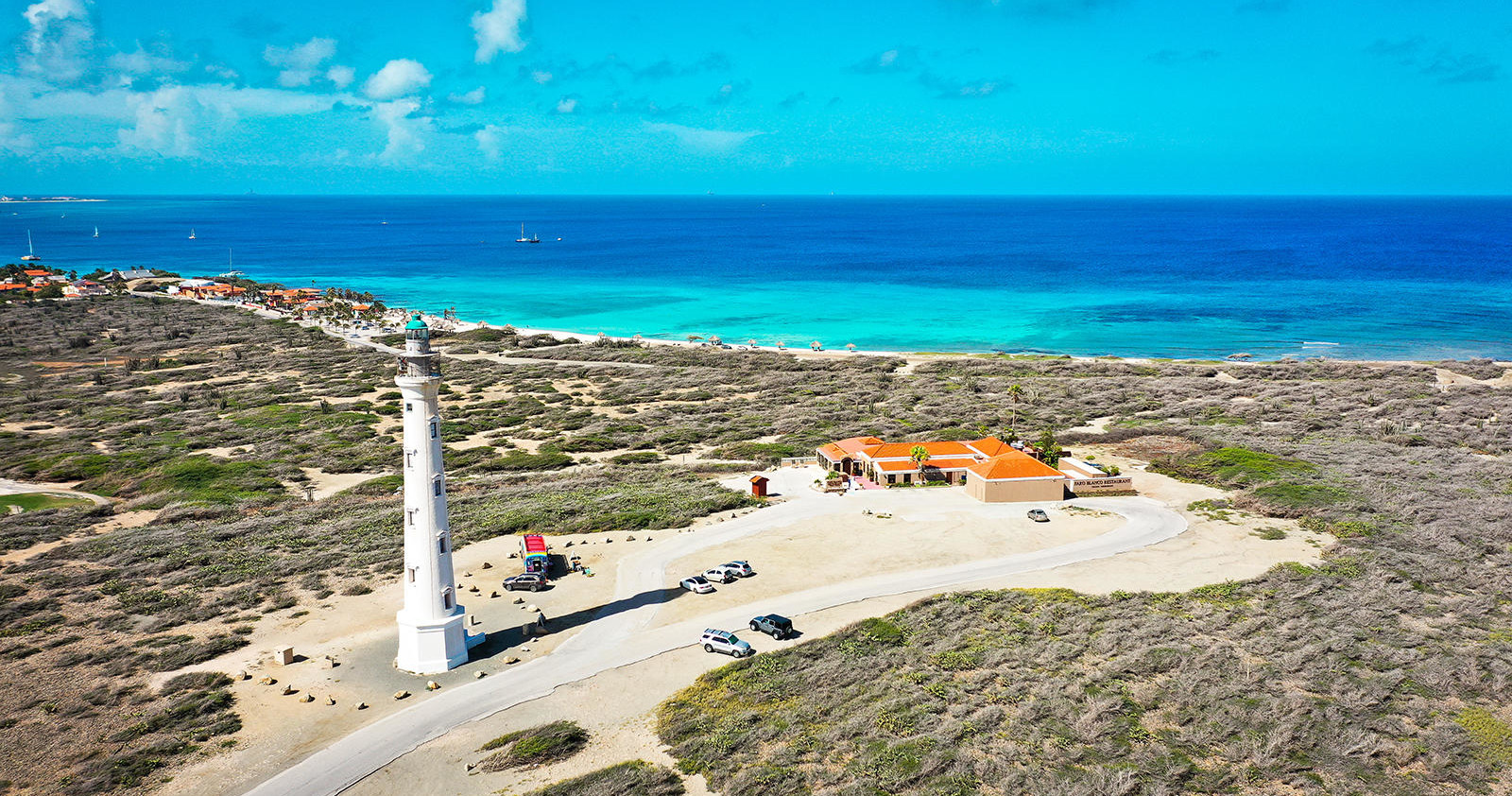 Tall lighthouse and building near coast with sandy landscape and clear blue sea.