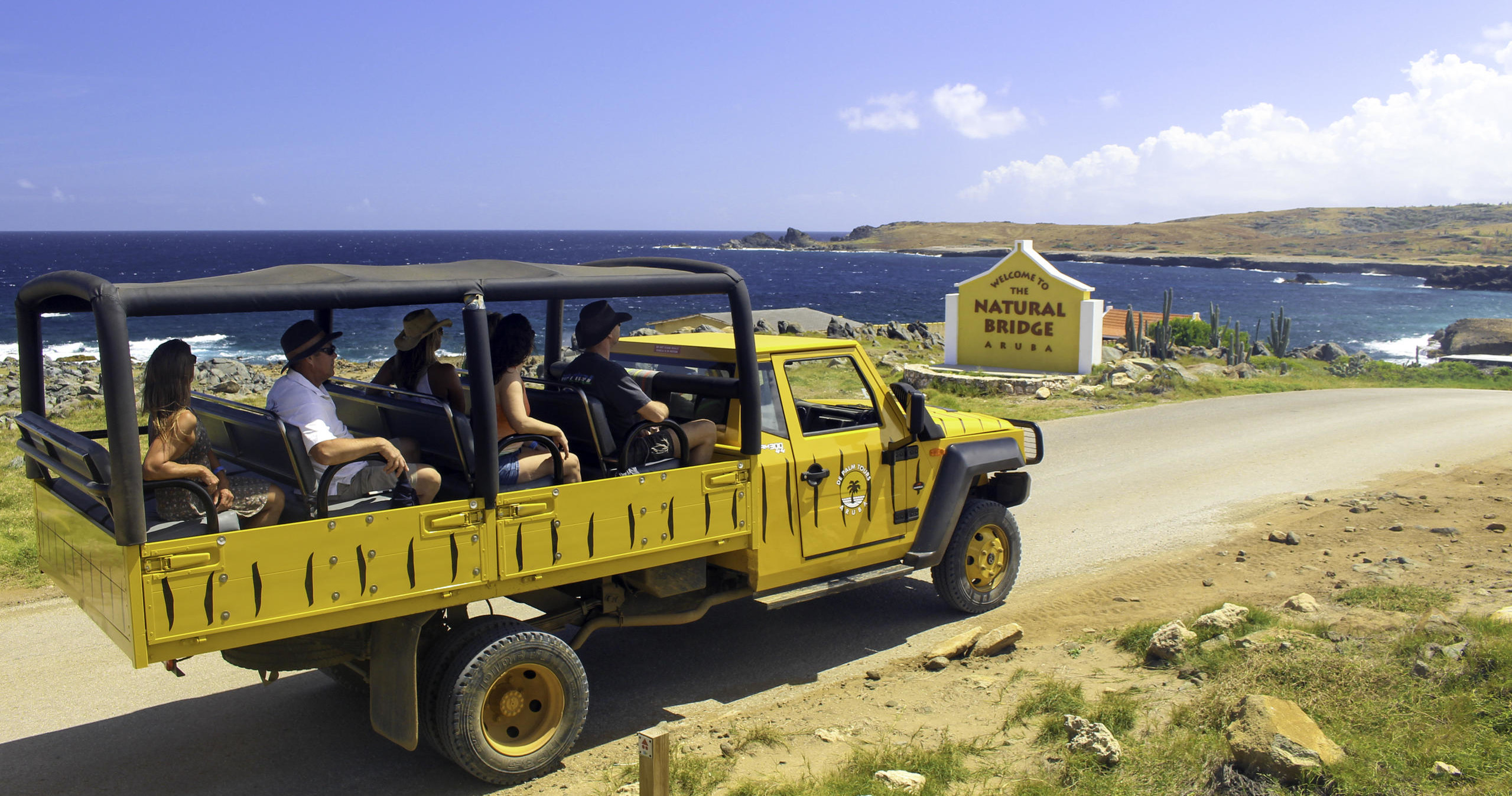 Open safari tour truck with passengers by coastline, near Natural Bridge sign.
