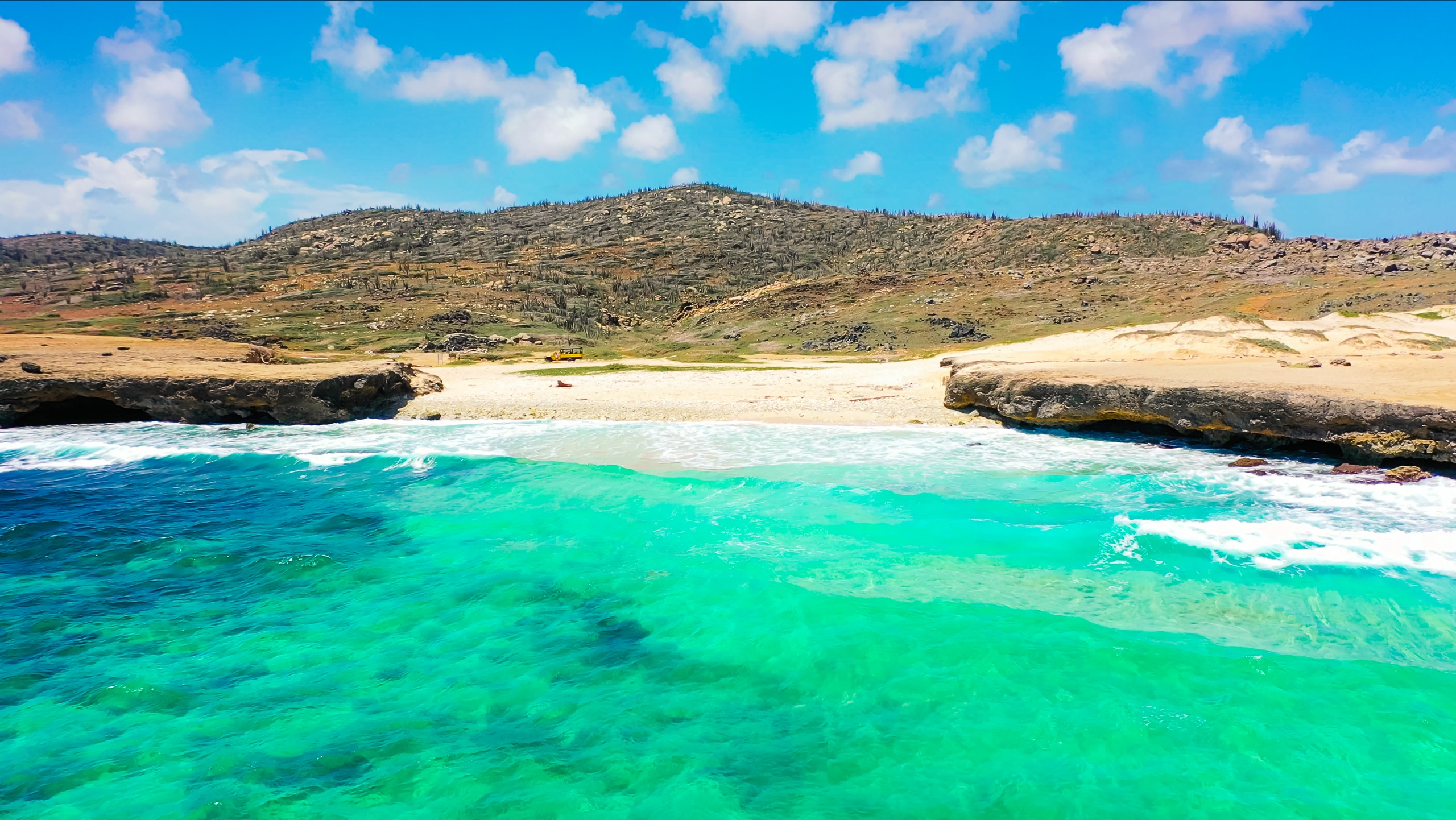 Turquoise sea with waves, rocky shore, and hills under a blue sky with clouds.