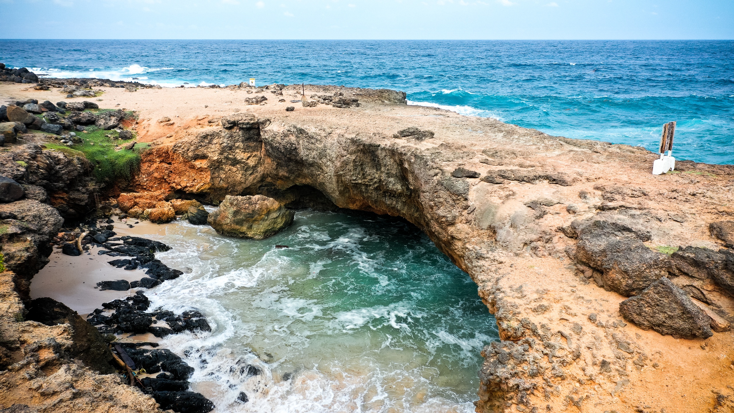 Rocky coastal arch with waves crashing underneath against a blue ocean backdrop.