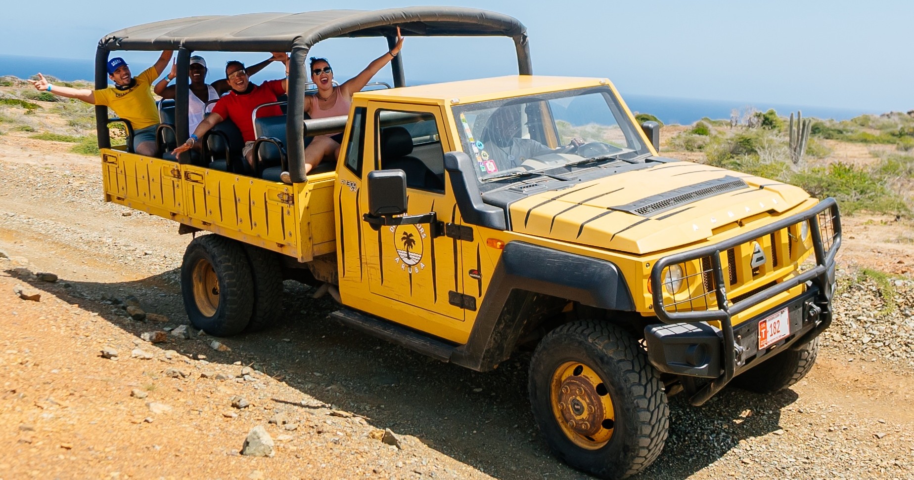 A yellow open-air jeep with passengers waving on a rocky coastal road.