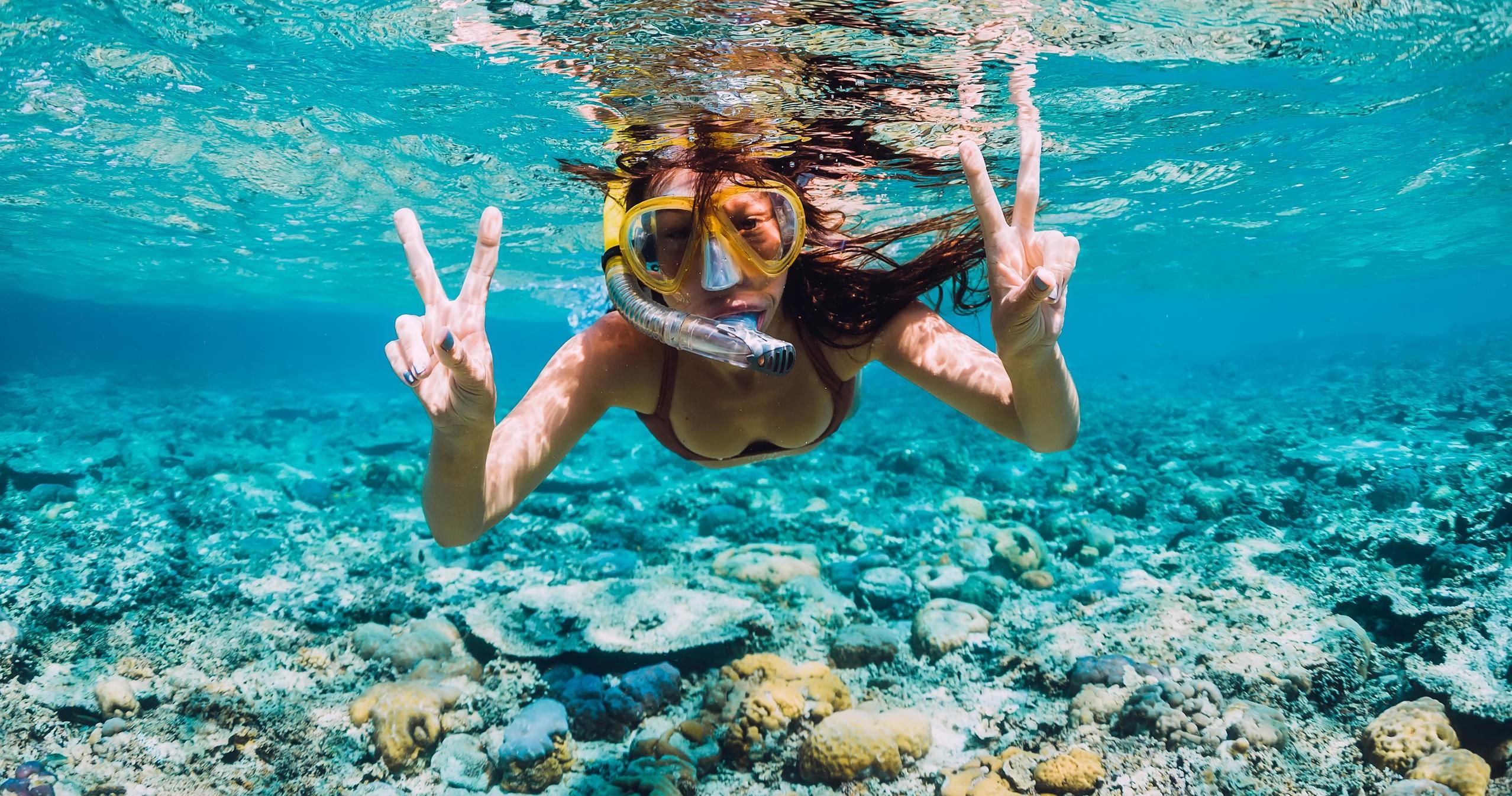 Person snorkeling underwater, showing peace signs with coral reef below.