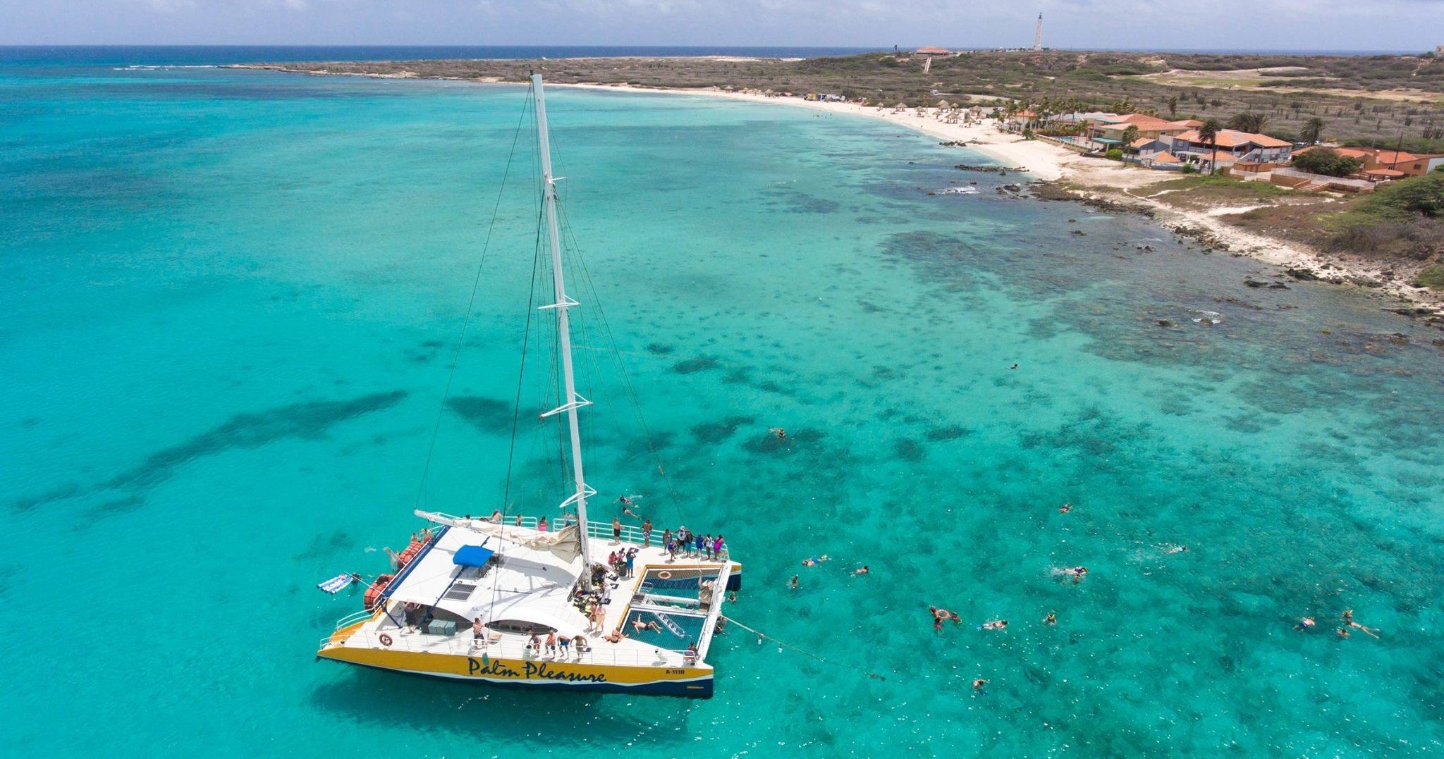 Yellow catamaran with people in turquoise water near a beach.