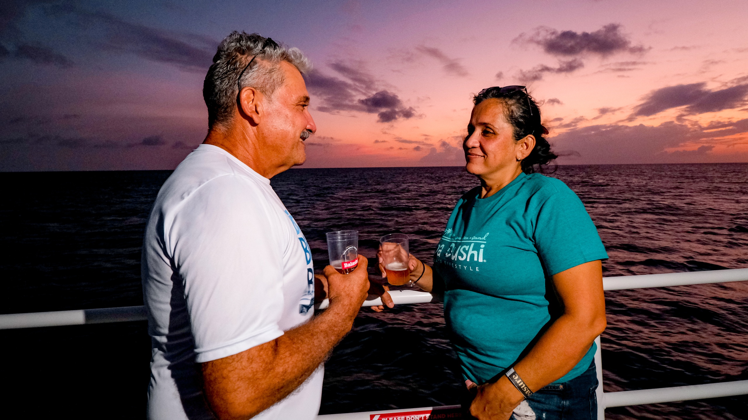Two people smiling and holding drinks on a boat at sunset.