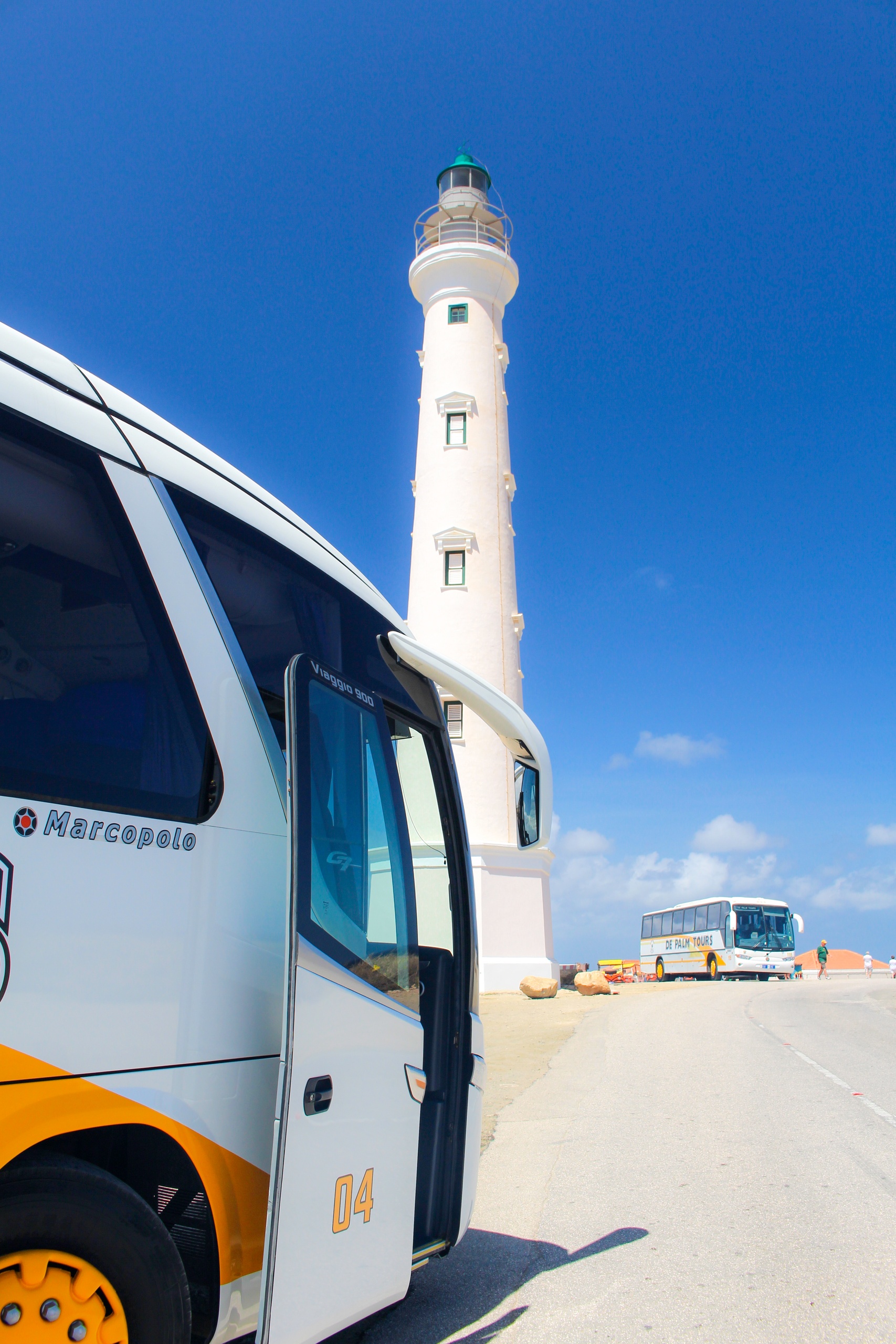 White lighthouse near two buses under a clear blue sky.