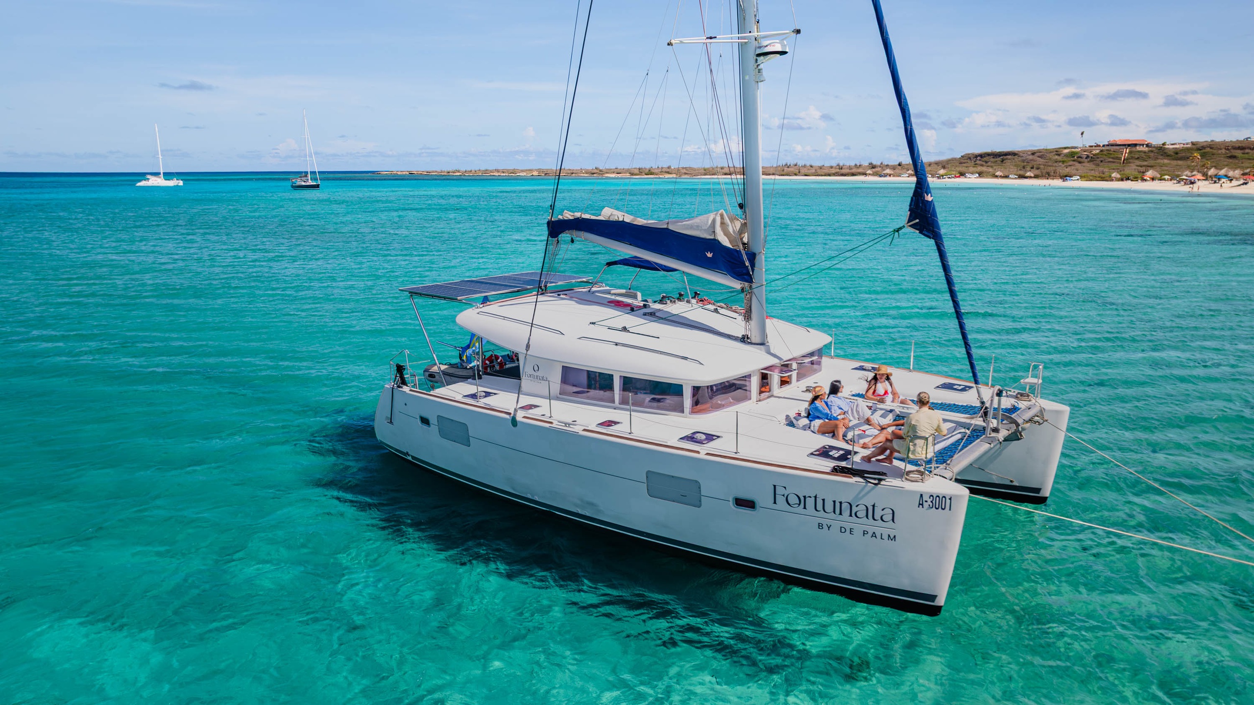 A catamaran sailing on clear turquoise water near a coastline with a sandy beach.