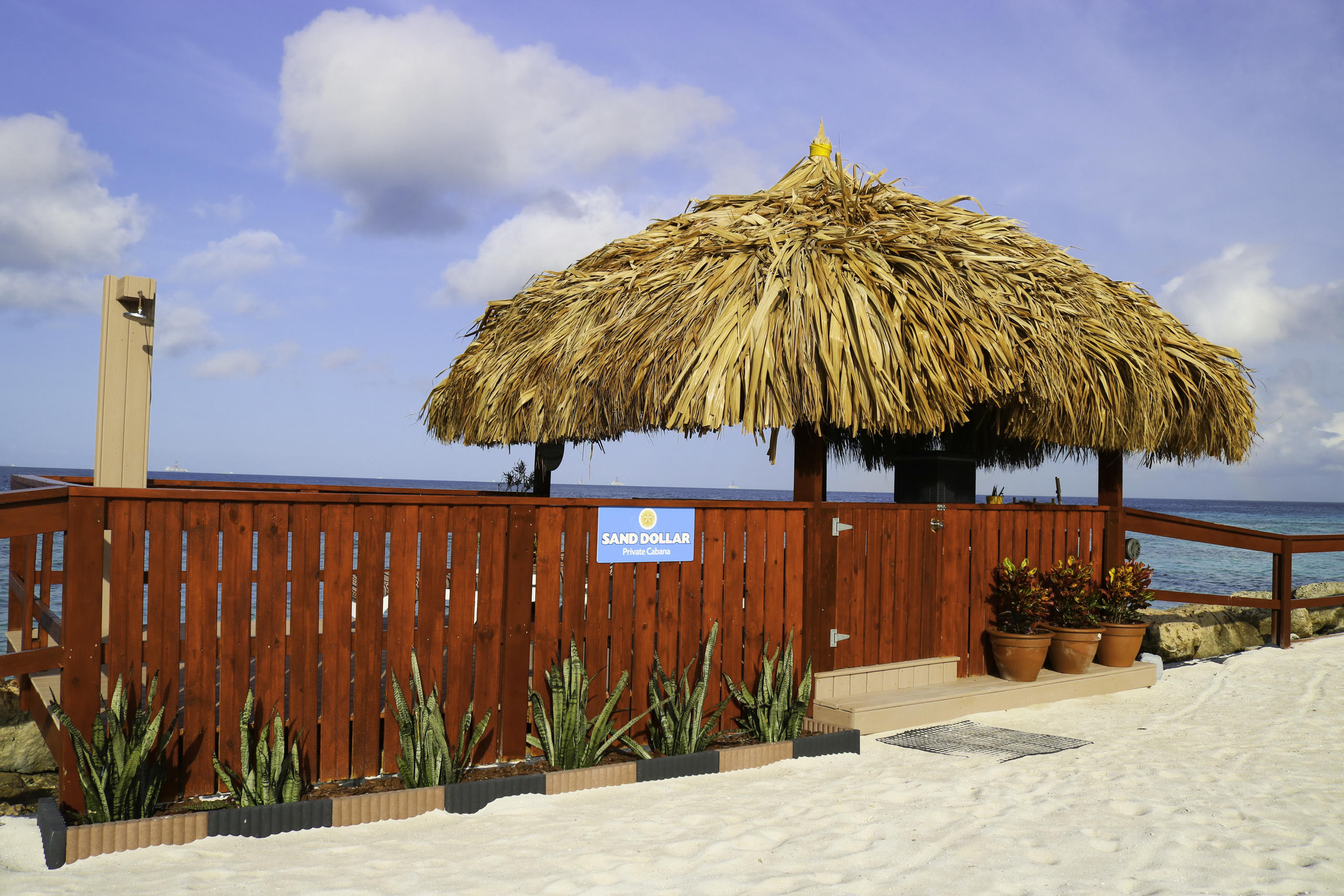 Beach hut with thatched roof, red fence, plants, and sand under a blue sky.