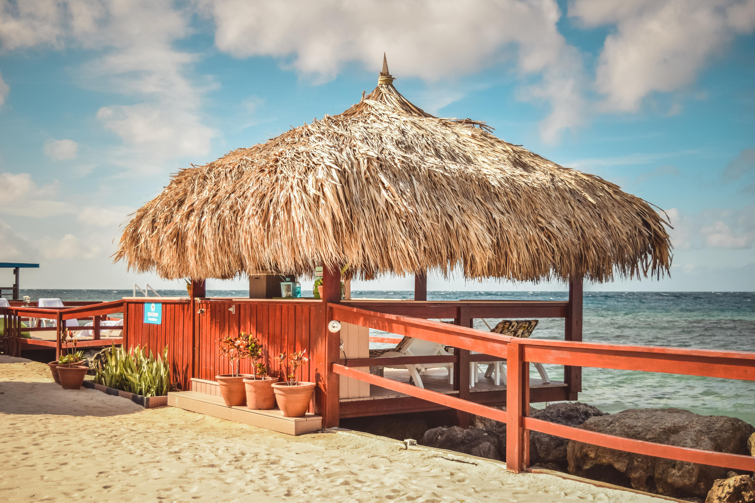 Beach hut with thatched roof, red railing, potted plants, ocean view, and blue sky.