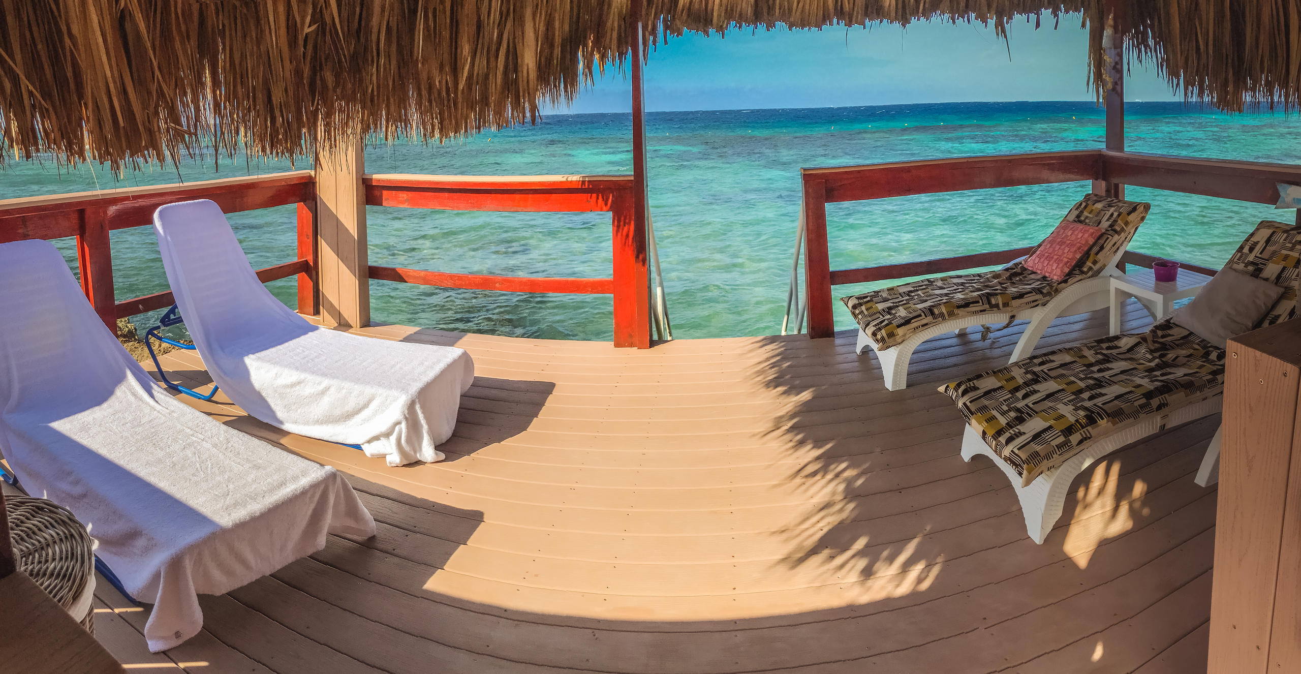 Deck with lounge chairs overlooking clear blue ocean under a thatched roof.