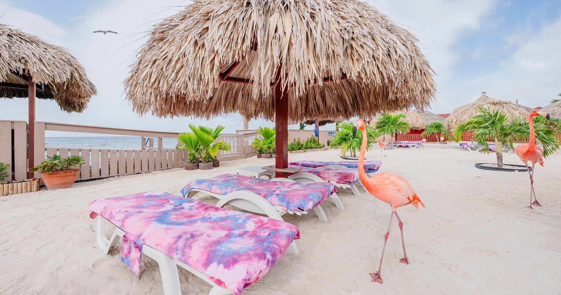 Beach scene with loungers, pink flamingos, and a palapa umbrella.