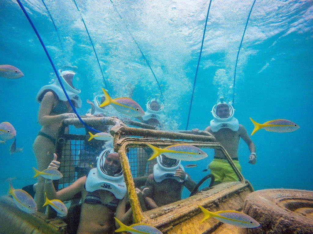 People underwater with helmets near a sunken vehicle surrounded by fish.