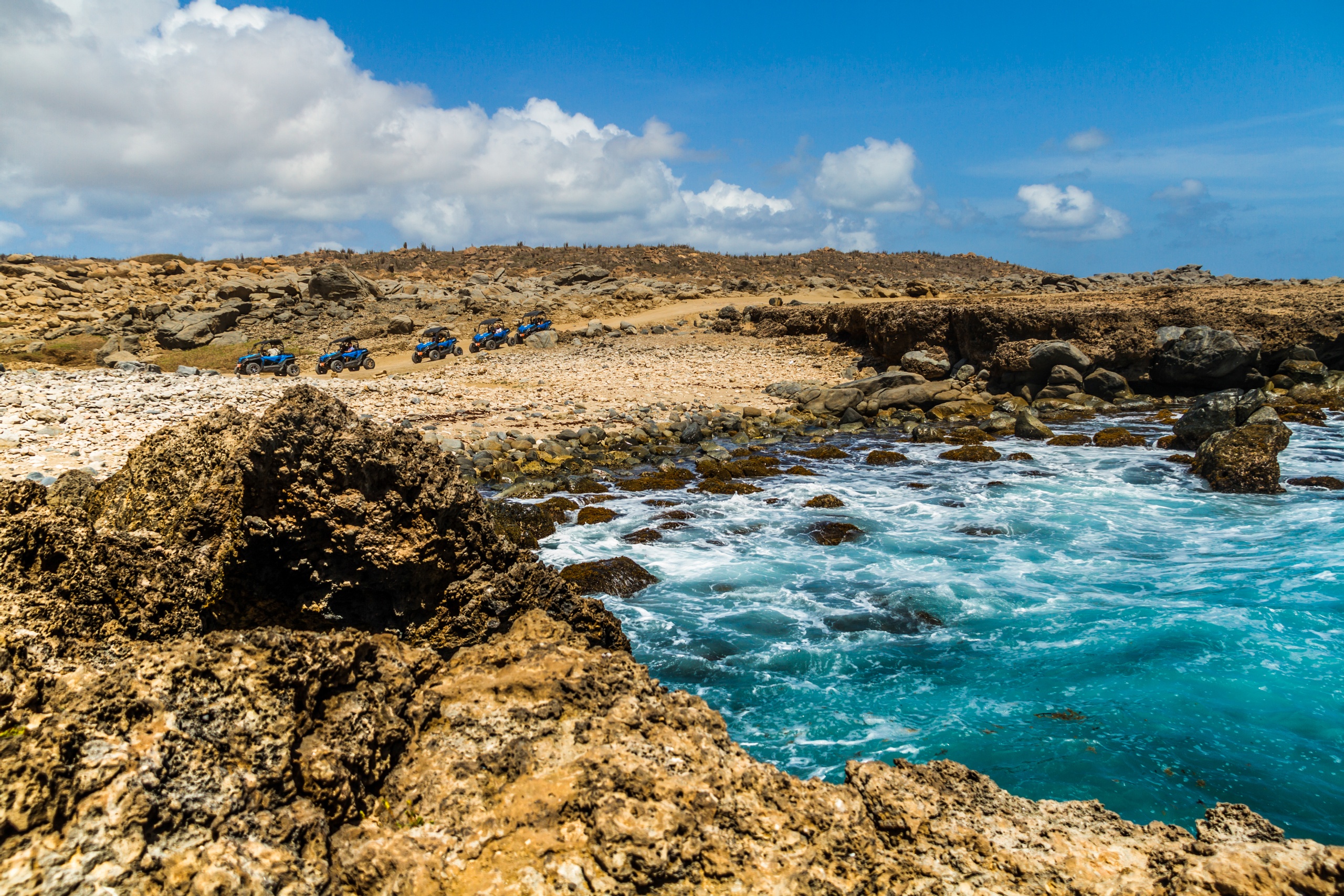 Rocky coast with blue ocean and row of off-road vehicles on a dirt path under a clear sky.