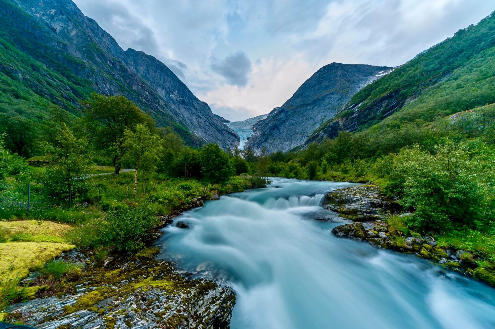 Mountain valley with a fast-flowing river and lush greenery under a cloudy sky.