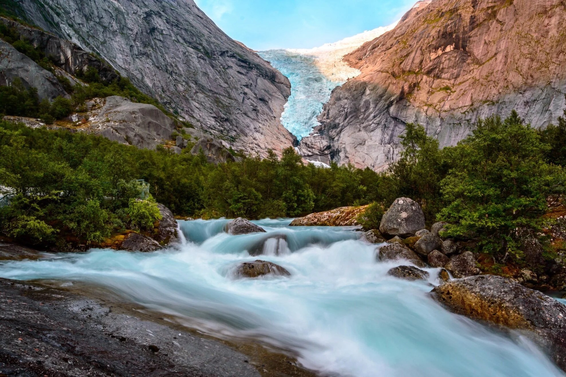 Mountain glacier with rushing river and lush greenery in the foreground.