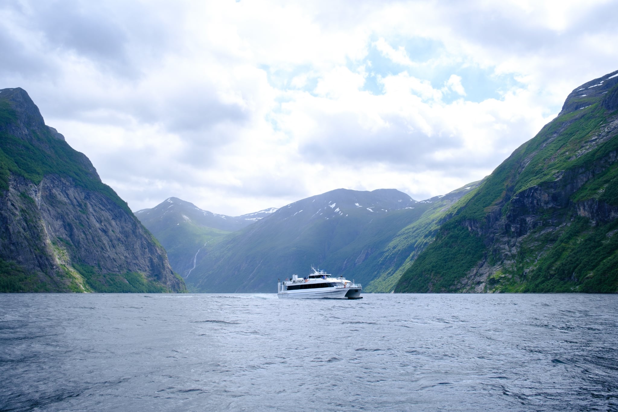 Ferry on water between steep, green mountains under a cloudy sky.