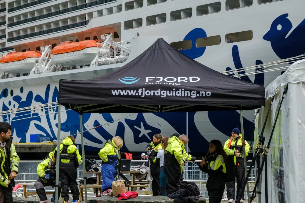 People in safety gear under Fjordguiding tent by a large ship.