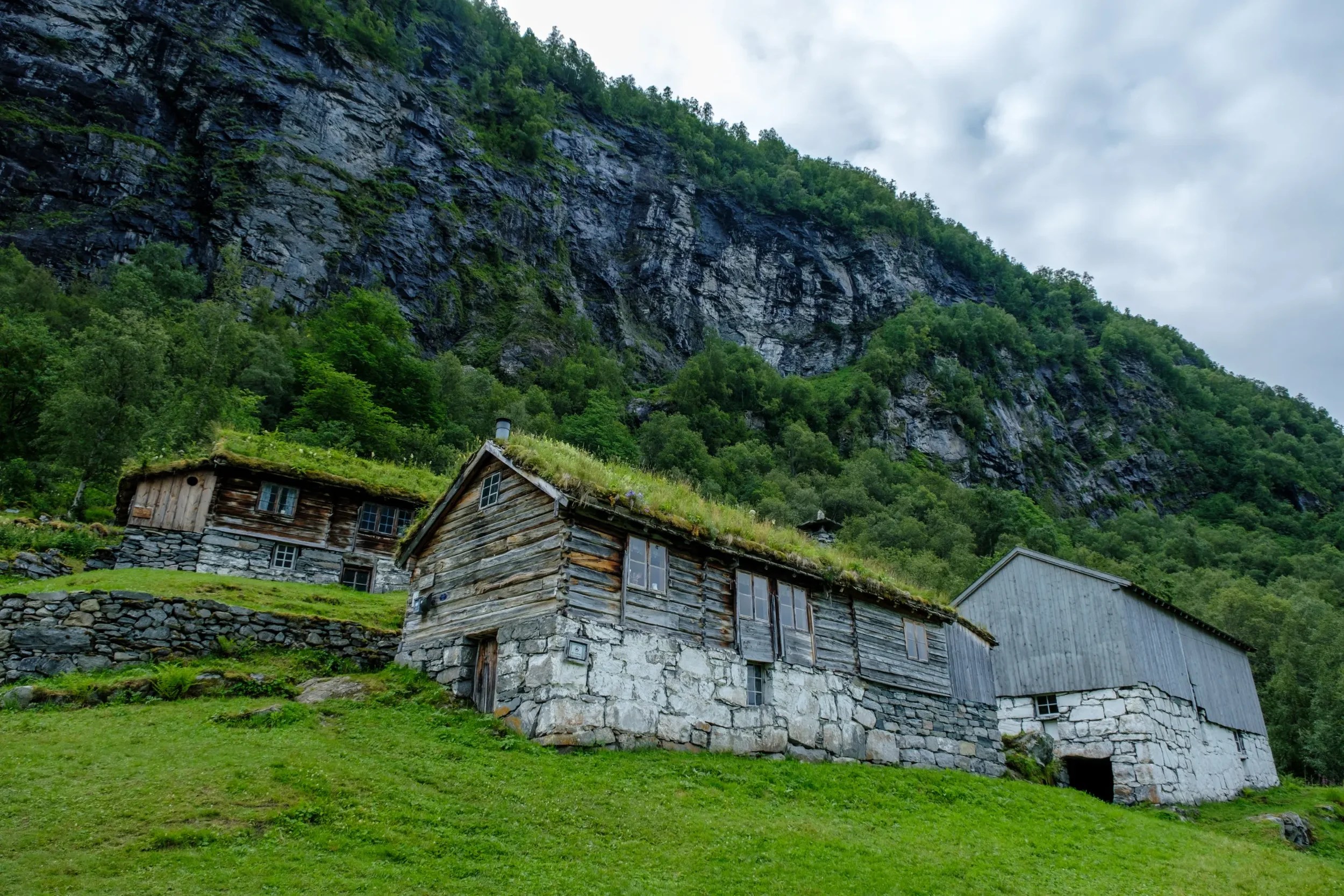 Rustic wooden cabins with grass roofs on a green hillside, beneath a rocky cliff and cloudy sky.