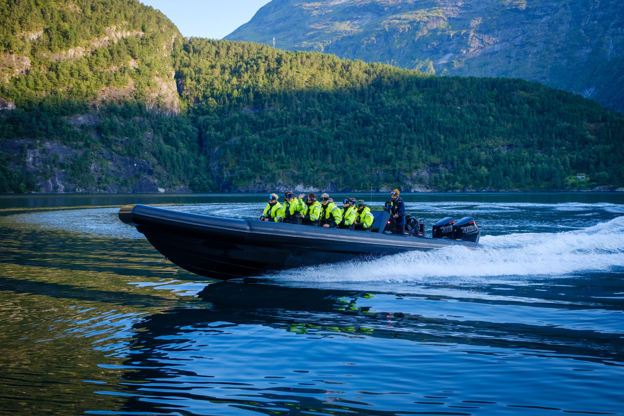Speedboat with passengers in safety gear travels on a scenic fjord.