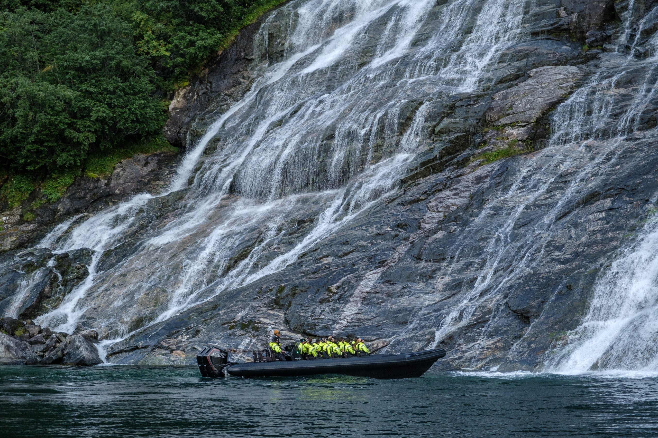 Tourists in yellow jackets on a boat near a large waterfall with rocky cliffs.