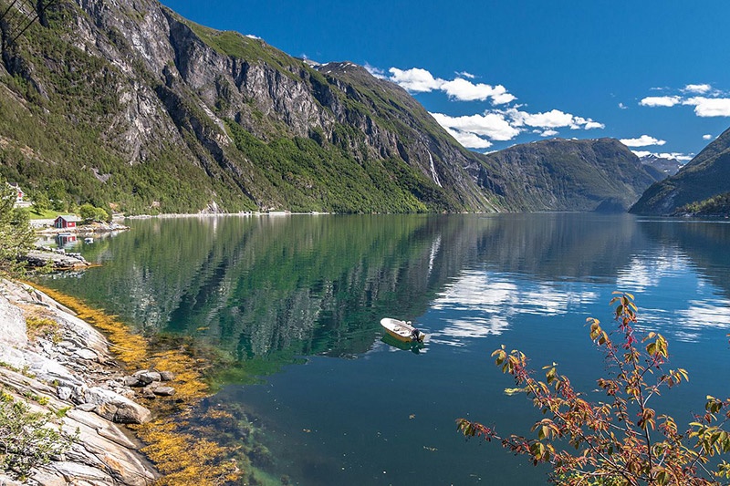 Tranquil fjord with mountains, blue sky, and small boat on the water.