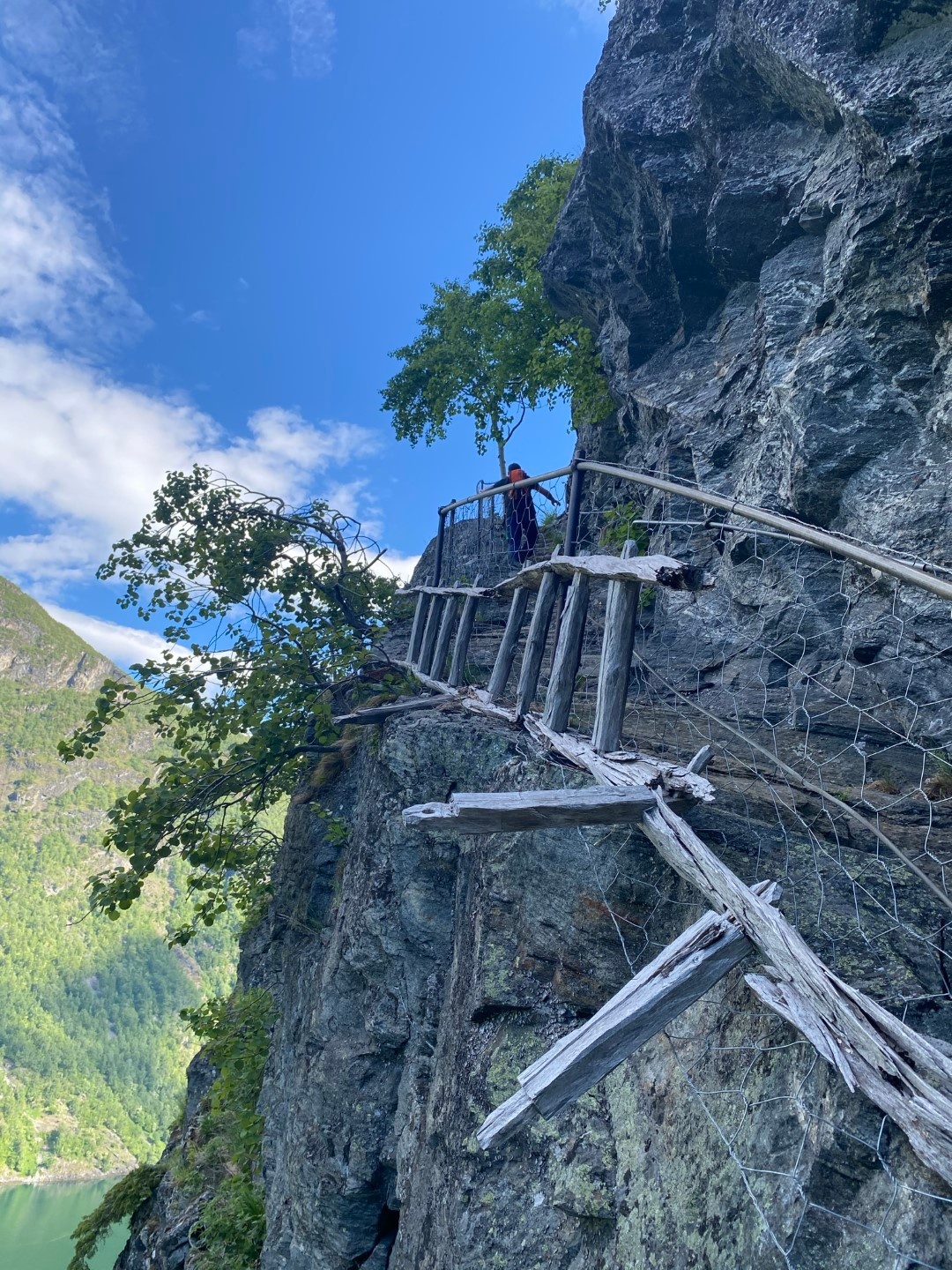 Person walking on narrow wooden path along a cliff with blue sky above.