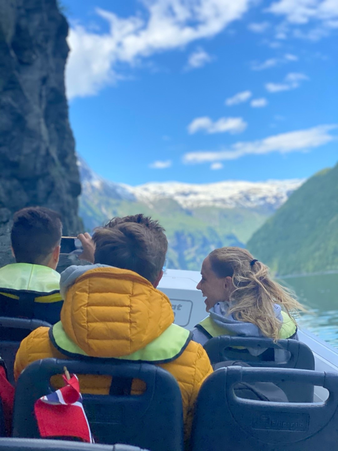 People in a boat with life jackets, surrounded by mountains and blue sky.