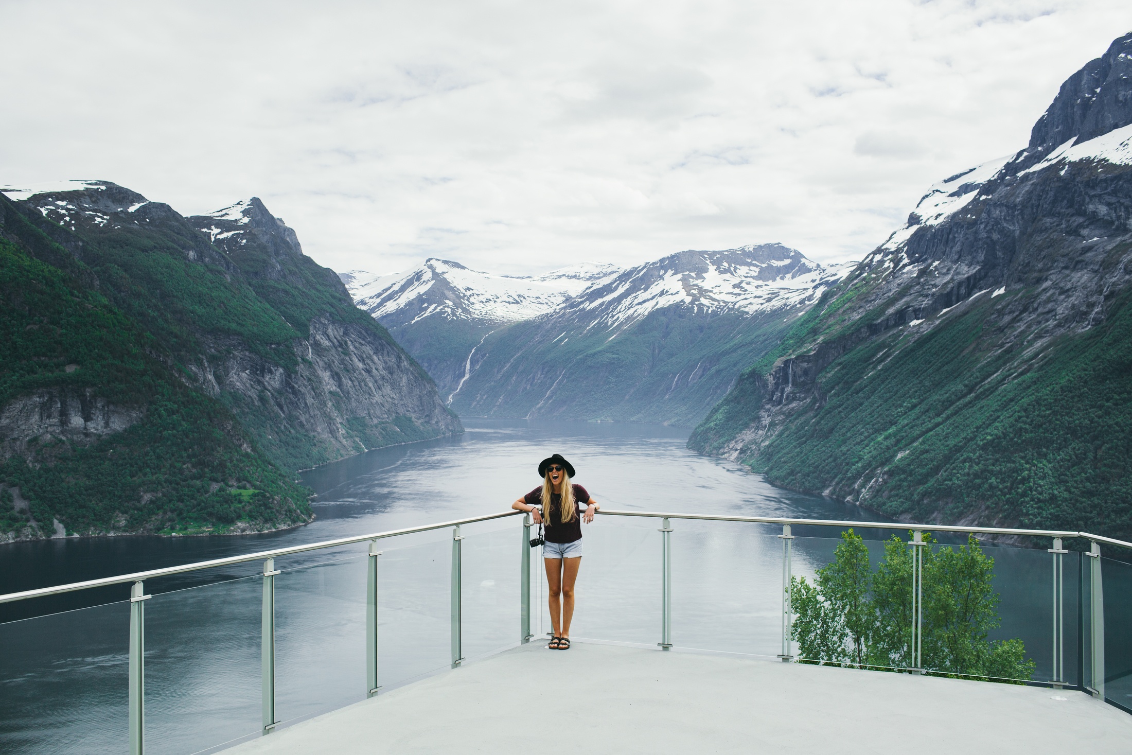 Person on balcony overlooking a fjord with mountains and cloudy sky.