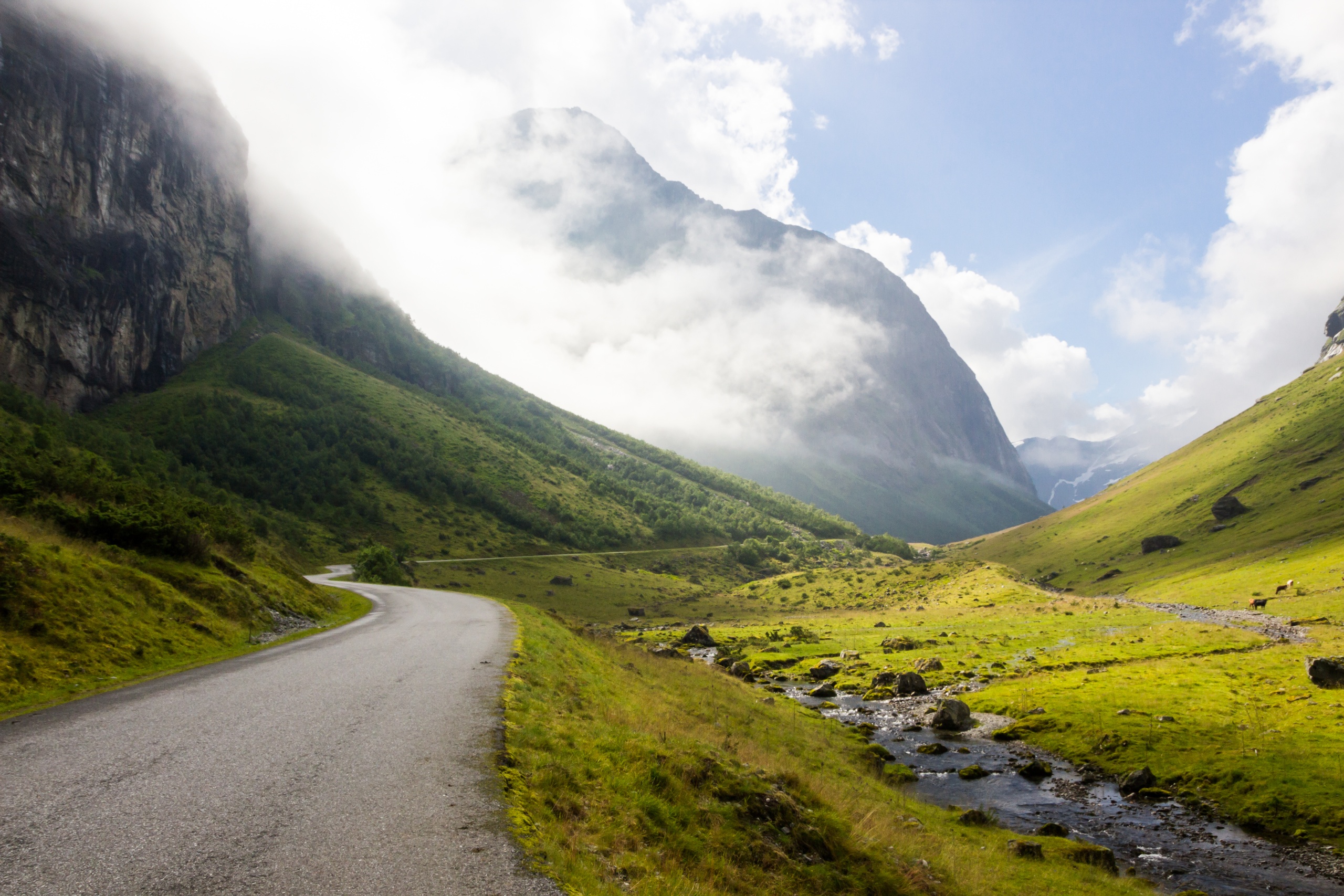 Mountain road with green hills, stream, and clouds.