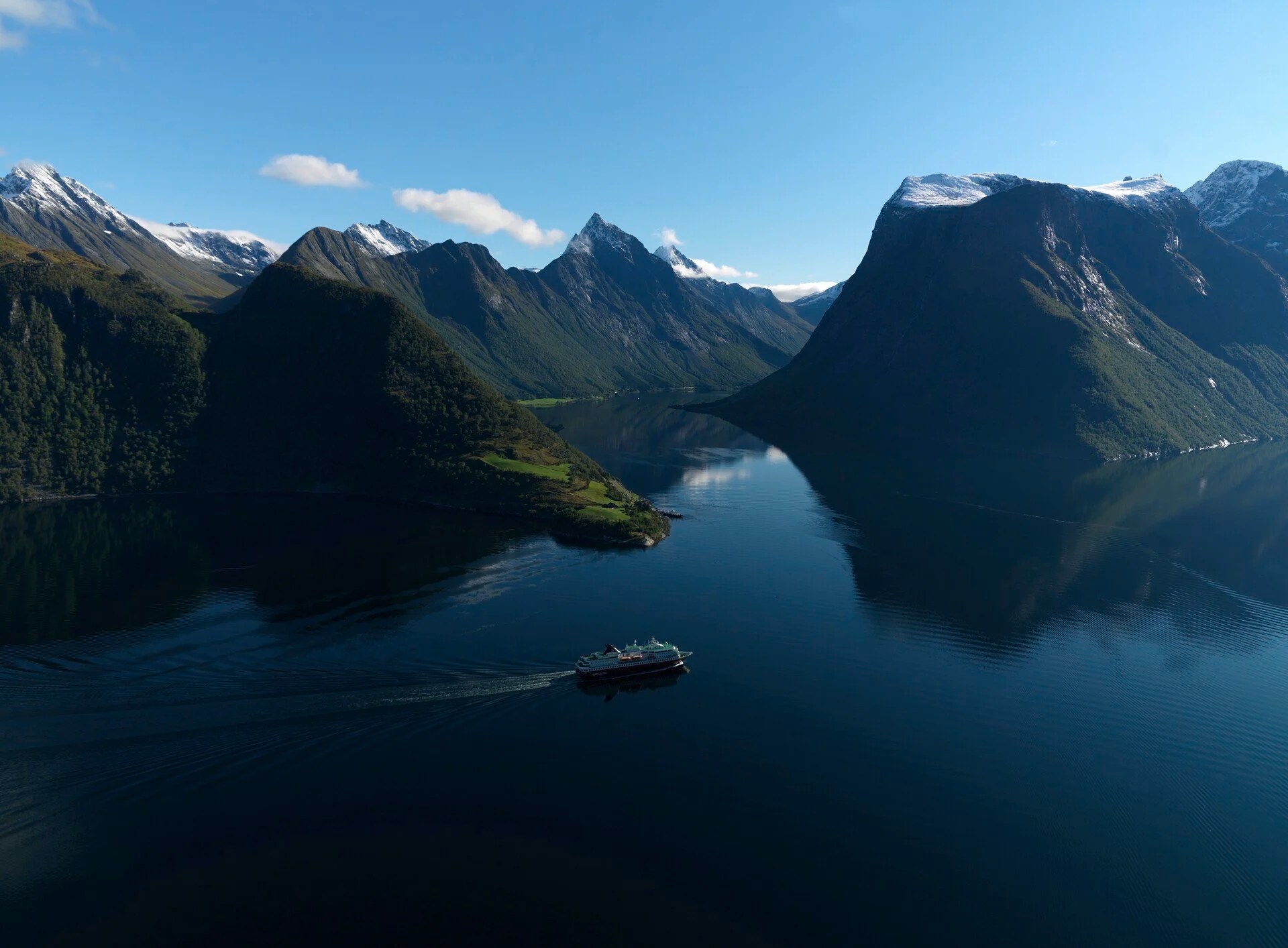 Aerial view of a ship sailing through a mountain fjord under a clear blue sky.