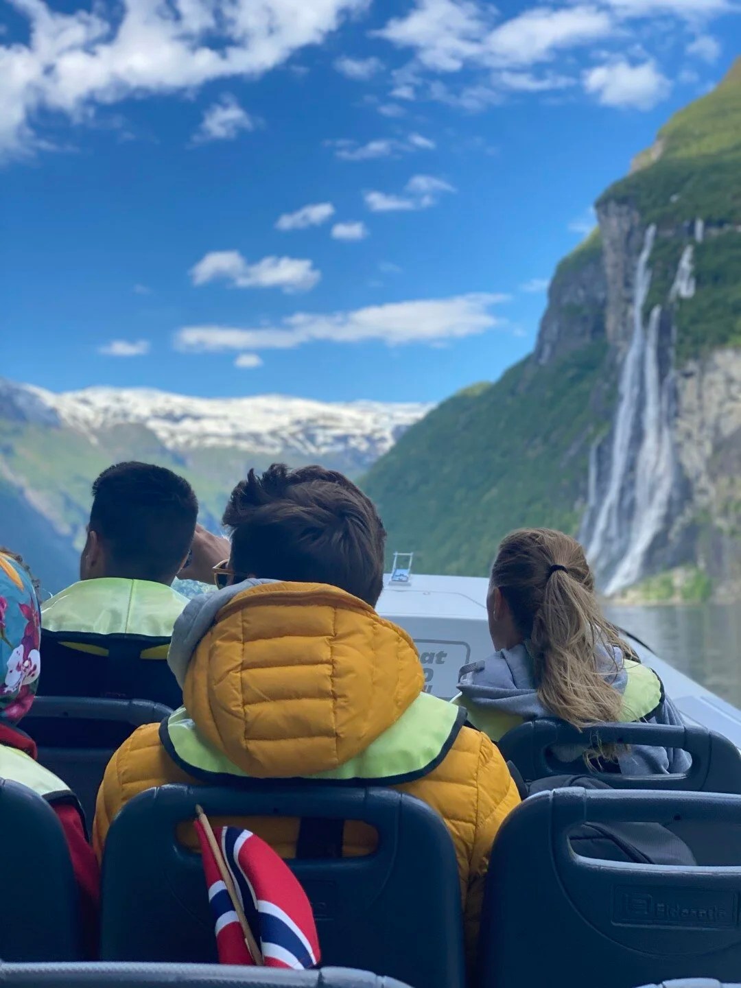 People on a boat view a waterfall and mountains under a blue sky.