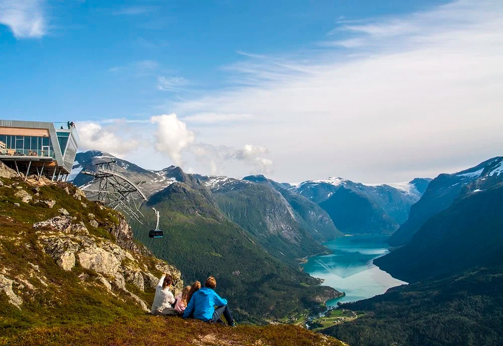 Three people sitting near mountain cabin overlooking lake and mountains.