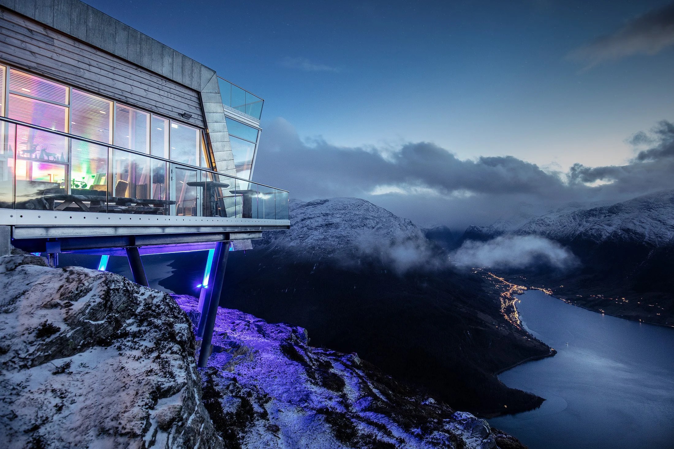 Modern building on cliff overlooking fjord and mountains at dusk.