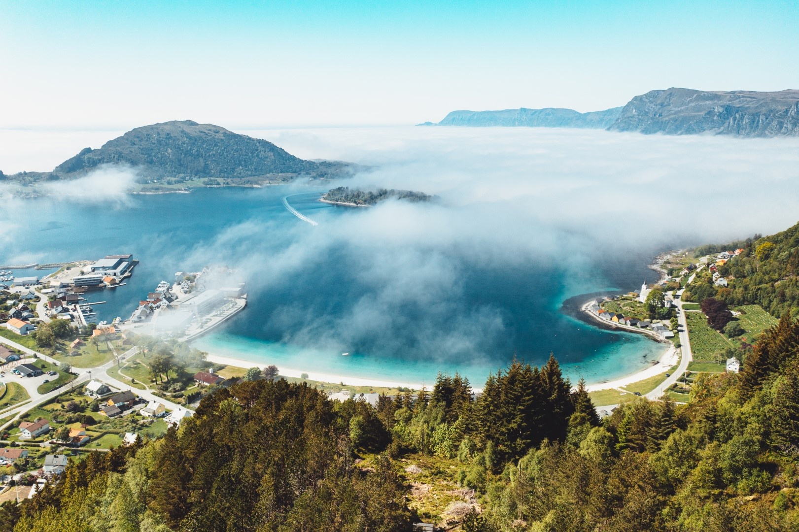 Aerial view of a coastal town, lush green hills, mist covering the bay, and distant mountains.