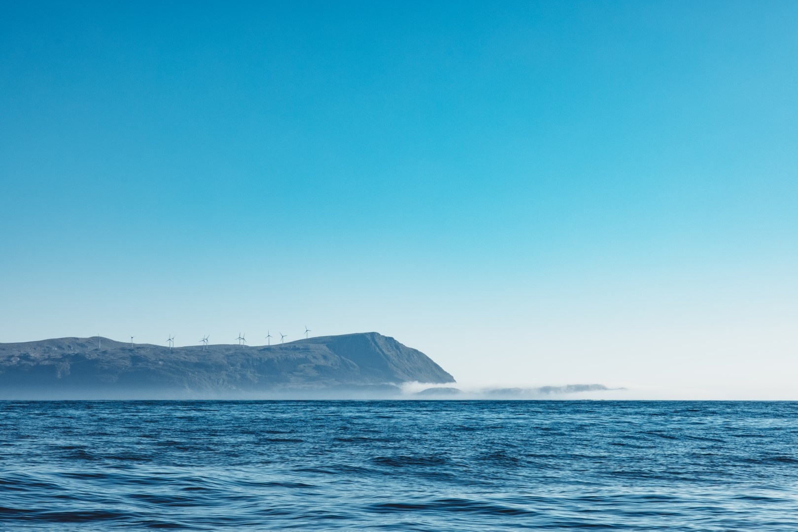 Ocean view with distant hills and wind turbines under a clear blue sky.