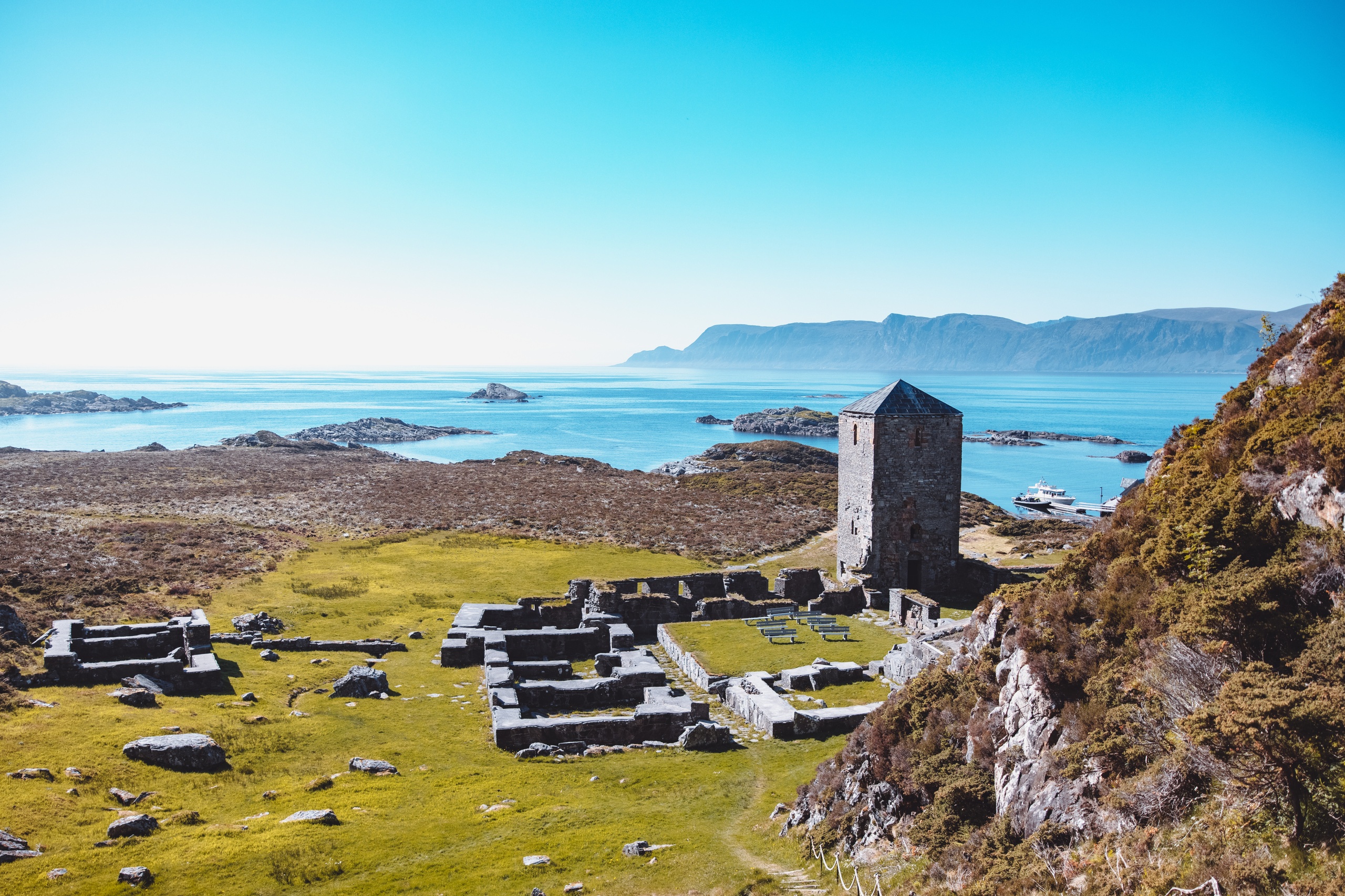 Ruins and tower by the sea with distant hills under a clear blue sky.