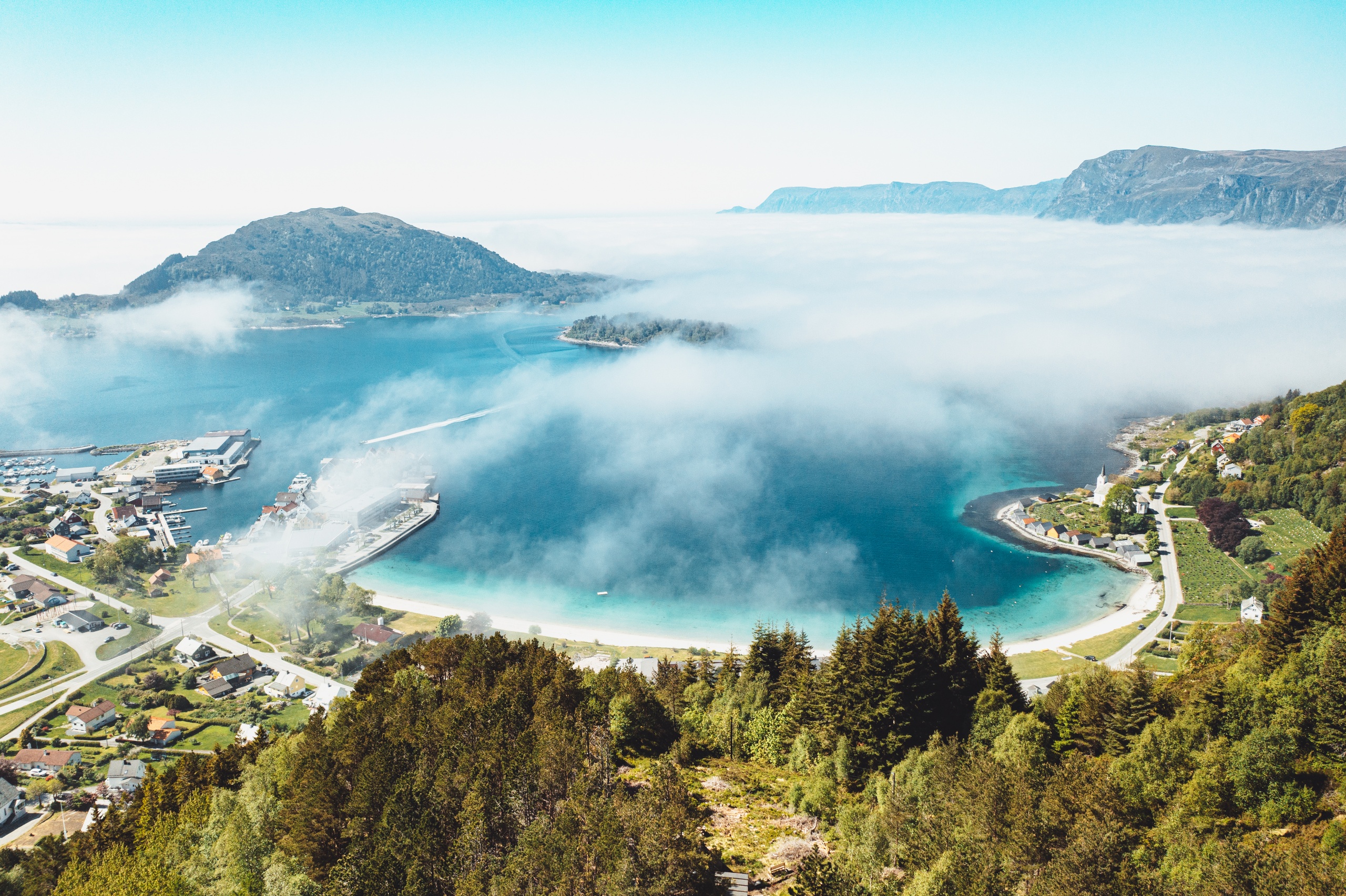 Aerial view of a coastal town by clear blue water, surrounded by misty hills and green forests.