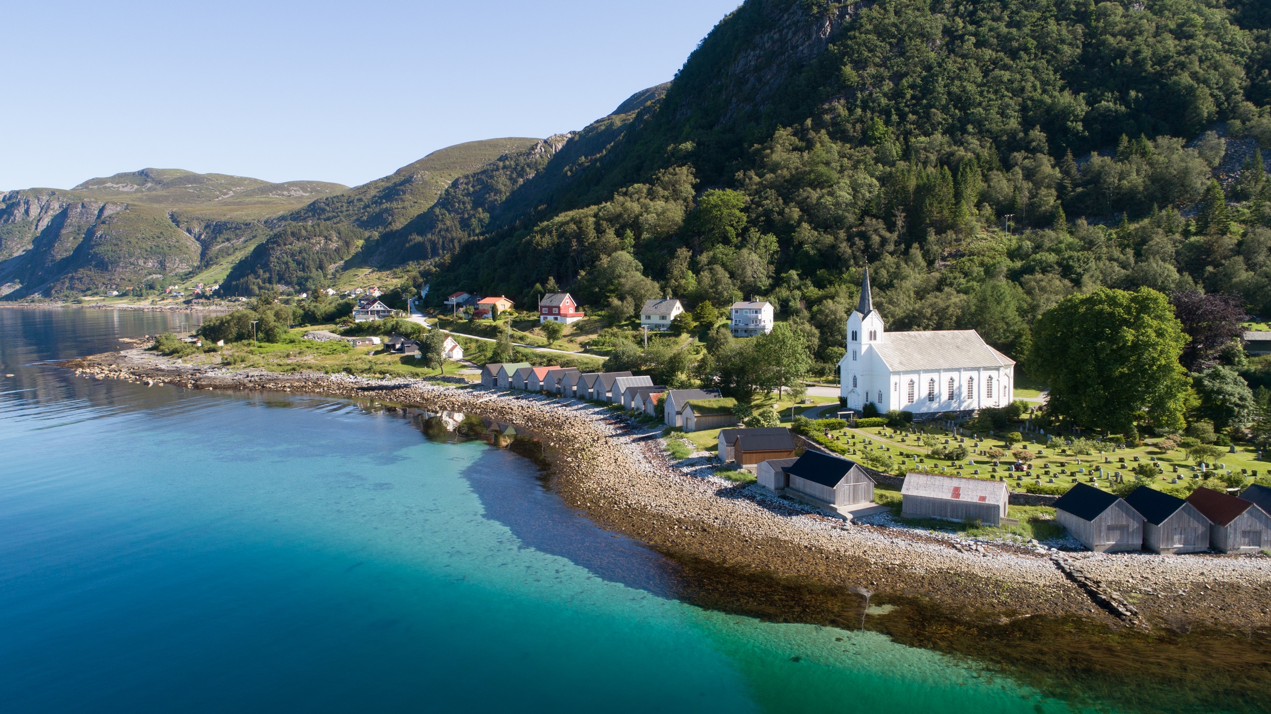 Coastal village with a white church beside a clear blue sea and surrounded by lush green hills.