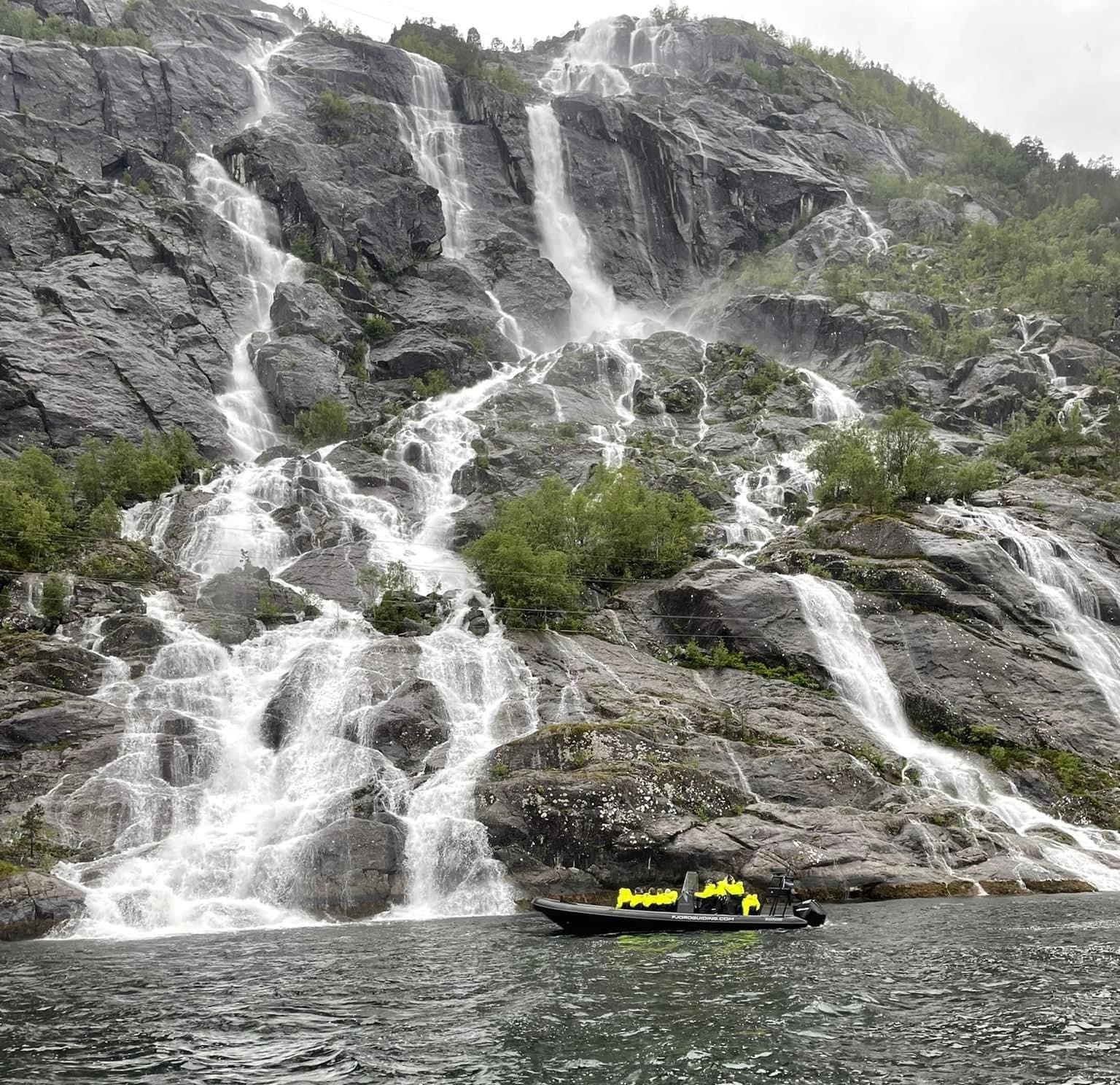 Inflatable boat near large waterfall on rocky cliff.
