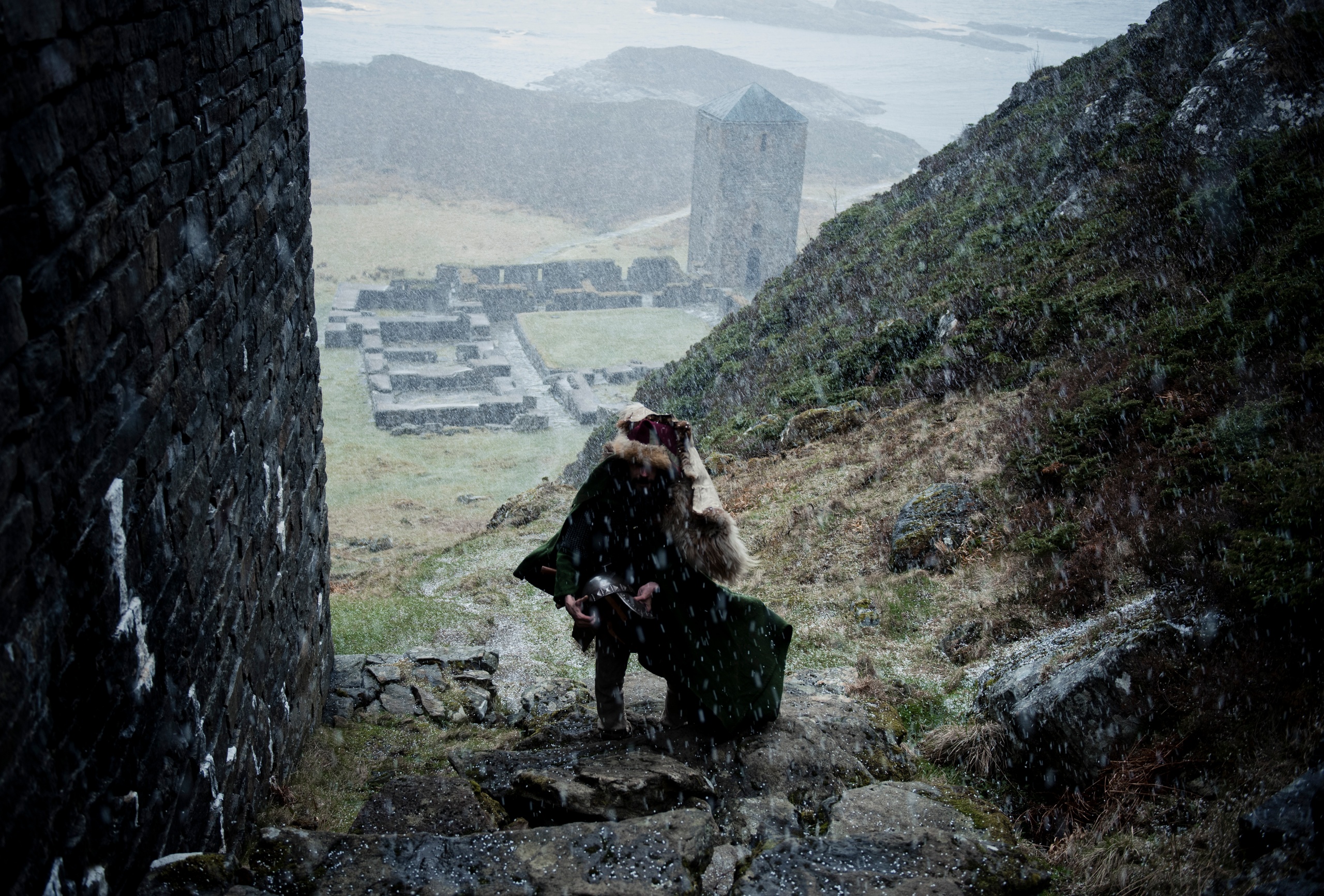 Person in cloak ascending stone steps in snow near ancient ruins and tower.