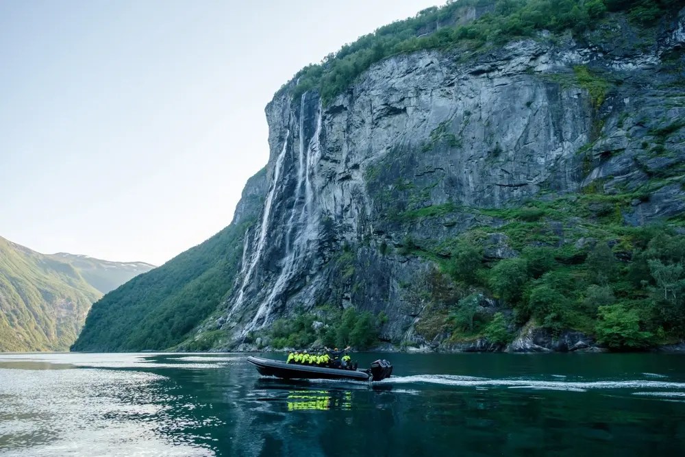 Boat on fjord with steep cliffs and waterfall in background.