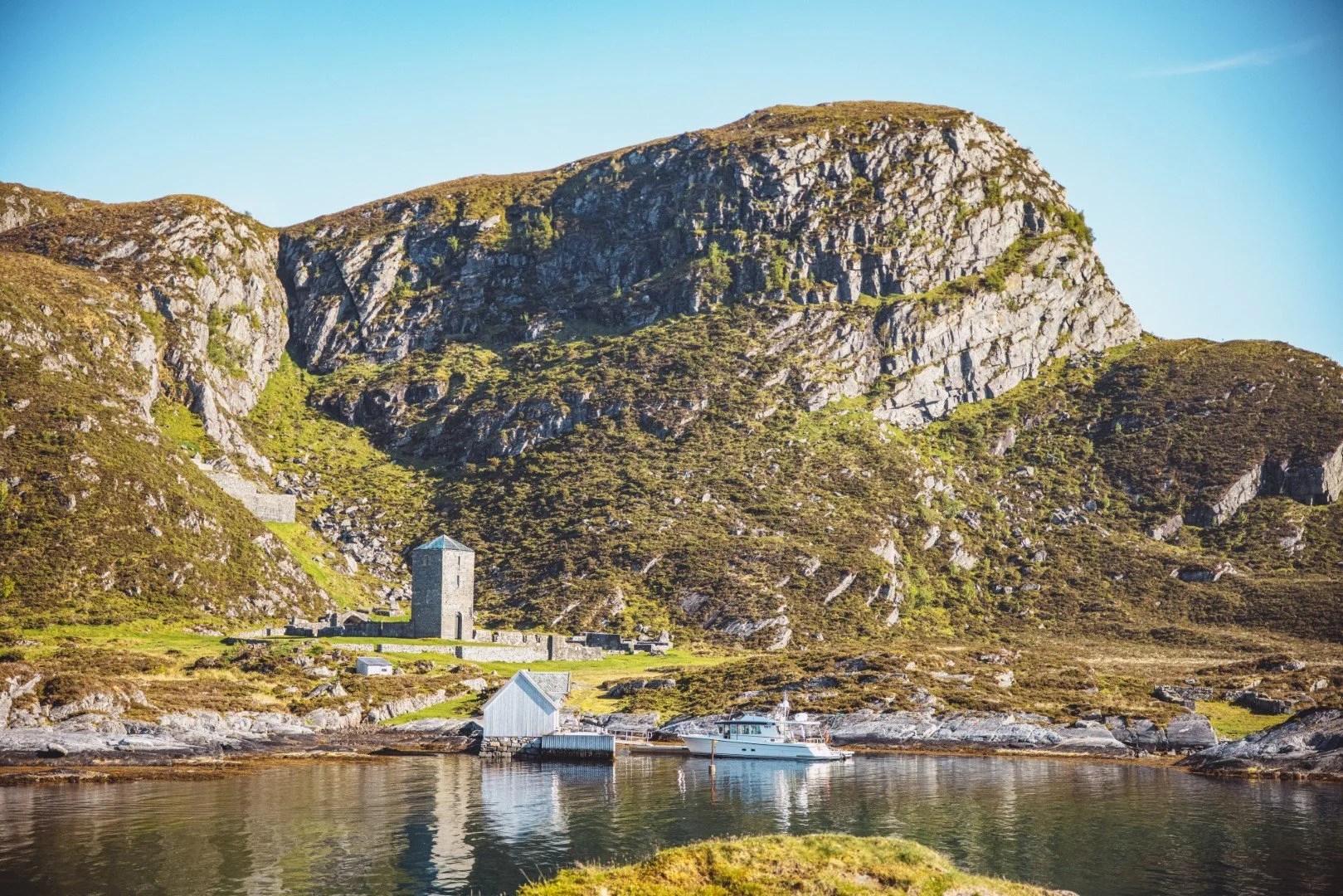 A rocky hillside with a tower, buildings, and a boat near the water.