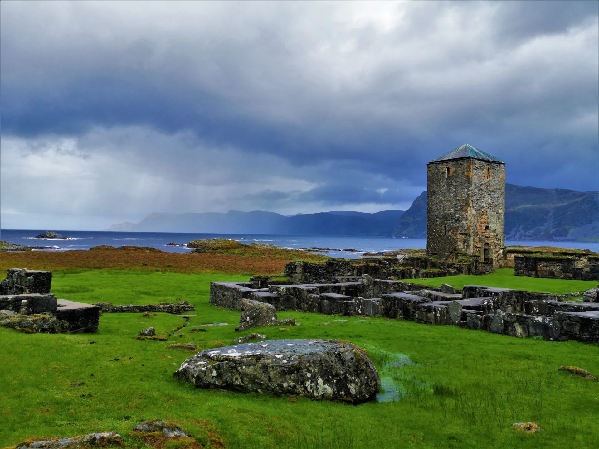 Ancient stone tower and ruins on a grassy shore with cloudy sky.