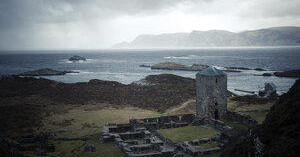 Old stone tower and ruins by a coastal landscape with mountains in the distance.