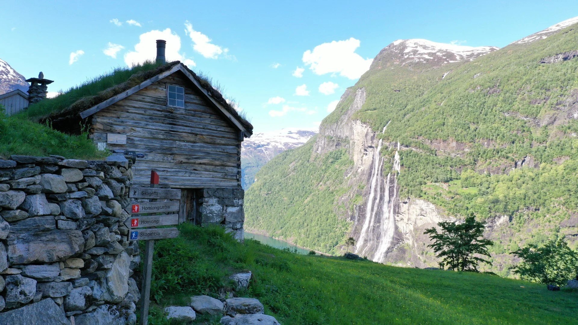 Wooden cabin with grass roof near waterfall and green mountains.