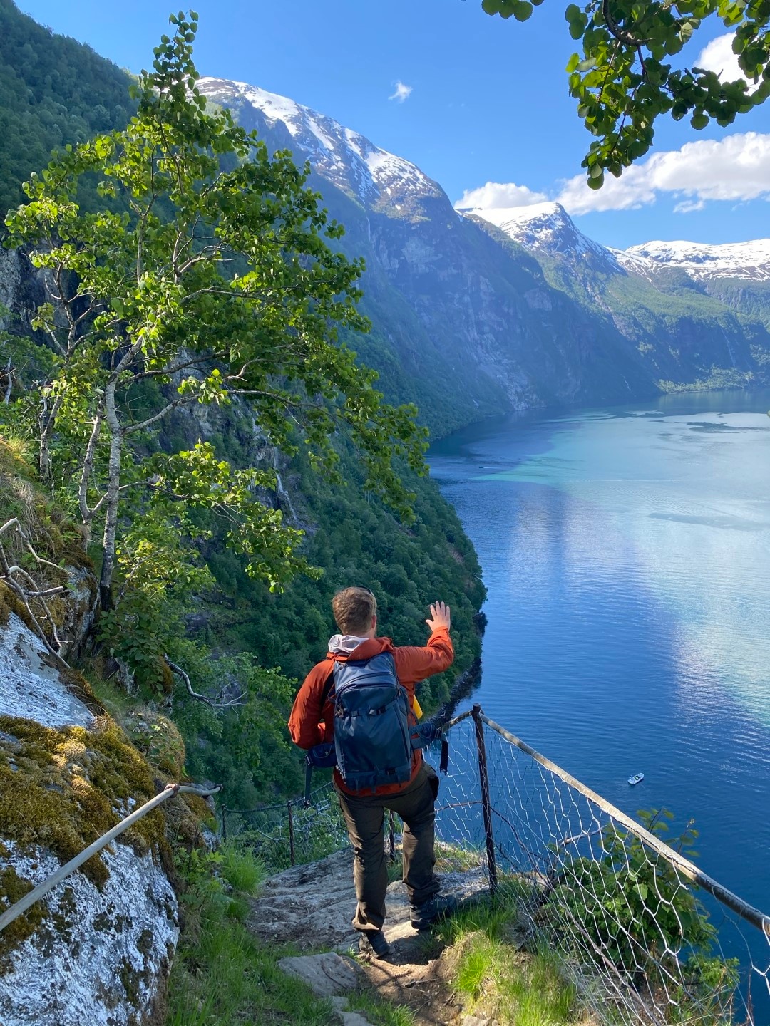 Person hiking on a mountain path overlooking a fjord under a blue sky.