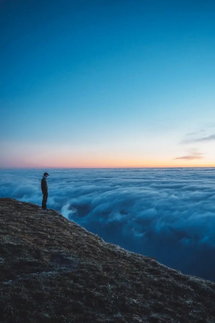 Person standing on a cliff edge above clouds at sunrise.